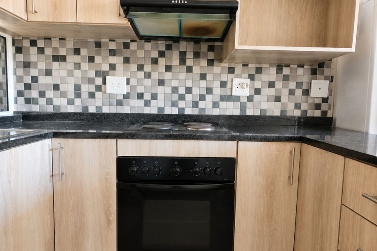 A functional kitchen featuring light wood-finish cabinetry, dark granite countertops, a mosaic tile backsplash, and integrated cooking appliances.