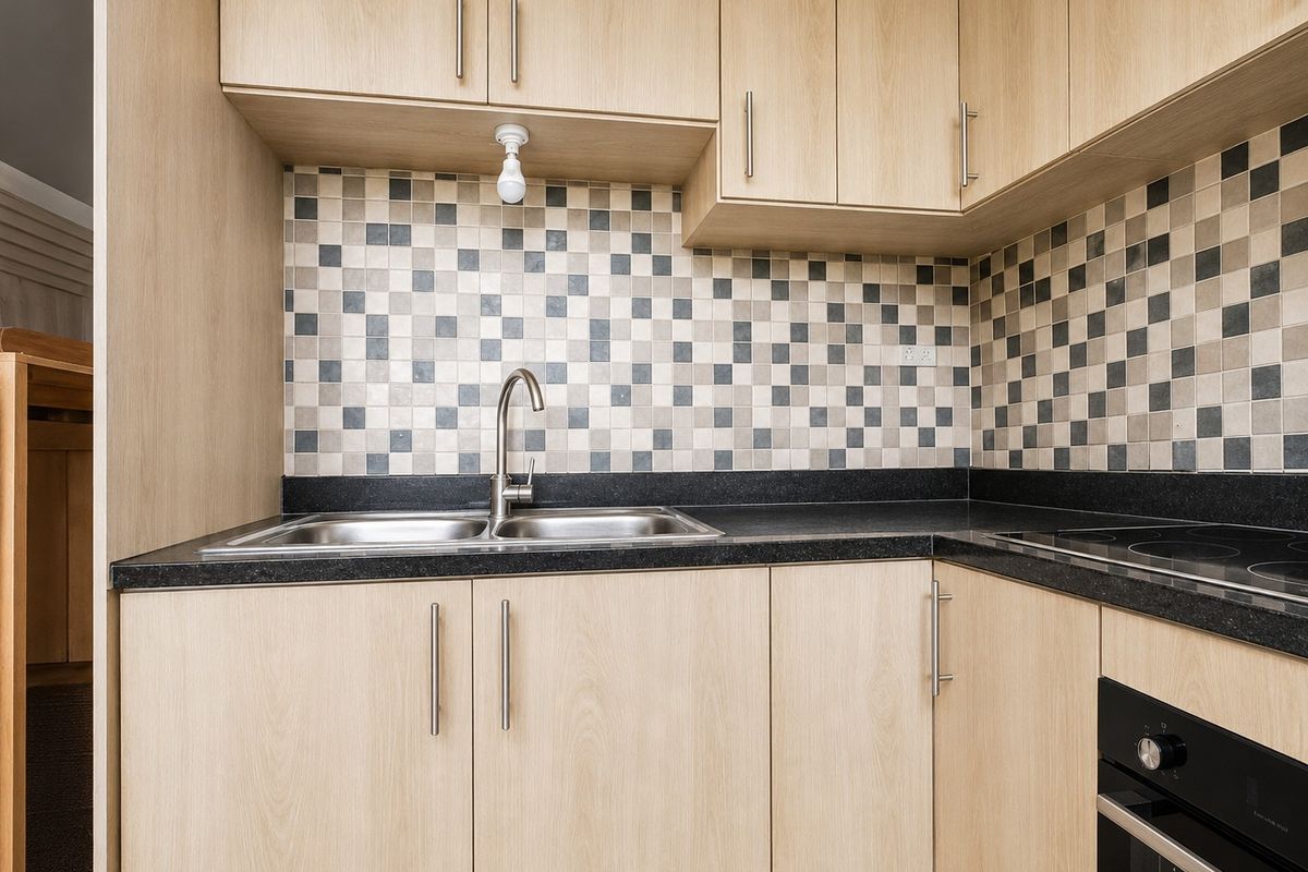 Compact kitchen featuring light wood-finish cabinetry, black granite countertops, a mosaic tile backsplash, and integrated stainless steel sink and oven appliances.