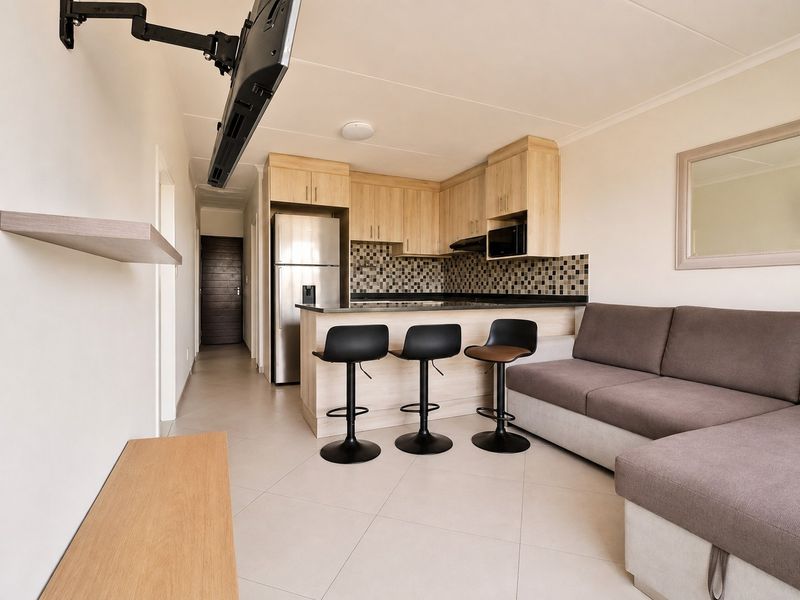 Open-plan living area featuring a compact kitchen with light wood cabinetry, mosaic backsplash, breakfast bar, and tiled flooring throughout.