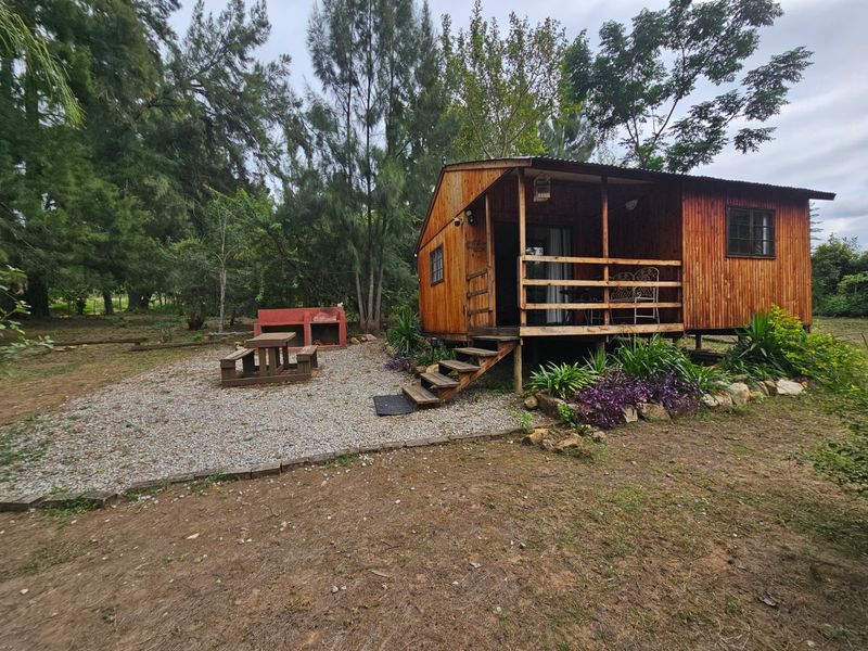 A rustic wooden cabin featuring a raised deck, gravel outdoor entertainment area, and a built-in brick braai set within a natural garden setting.