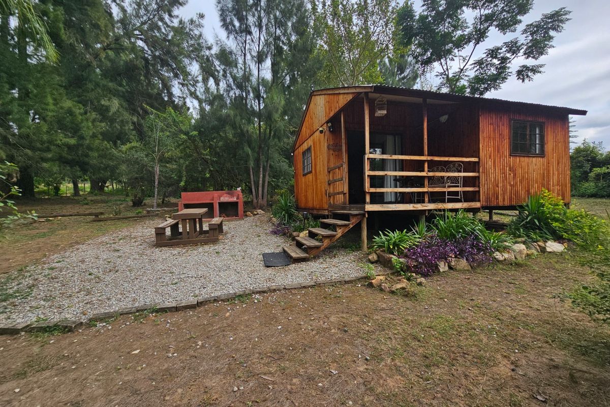 A rustic wooden cabin featuring a raised deck, gravel outdoor entertainment area, and a built-in brick braai set within a natural garden setting.
