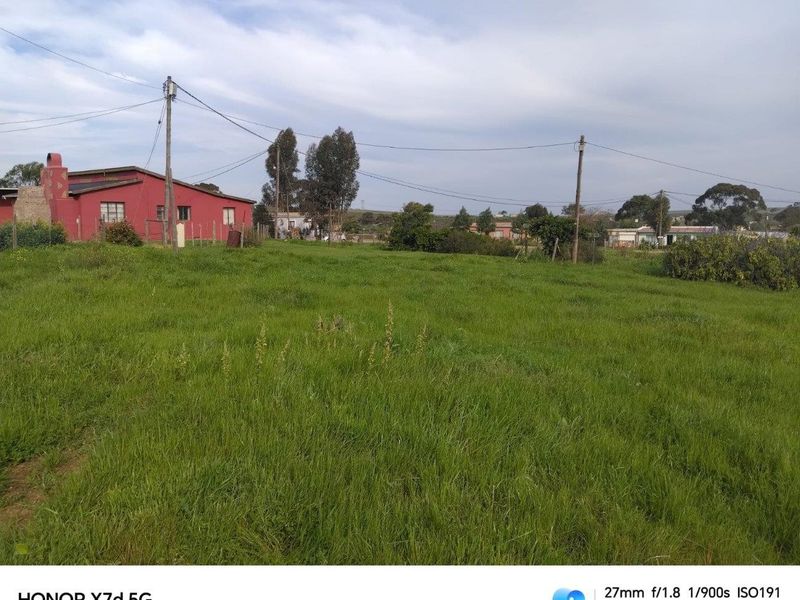 A view of a vacant residential plot featuring grassy terrain, utility poles, and overhead power lines with neighboring structures visible in the background.