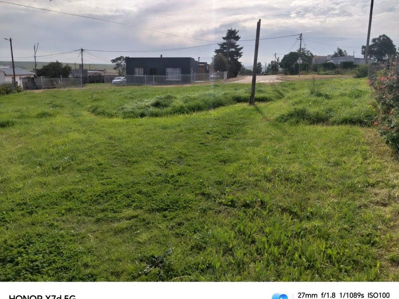 A vacant residential plot featuring grass coverage, perimeter fencing, and visible utility infrastructure, situated in a suburban street setting.