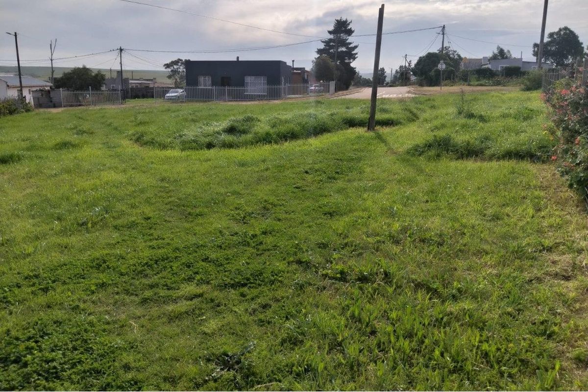 A vacant residential plot featuring grass coverage, perimeter fencing, and visible utility infrastructure, situated in a suburban street setting.