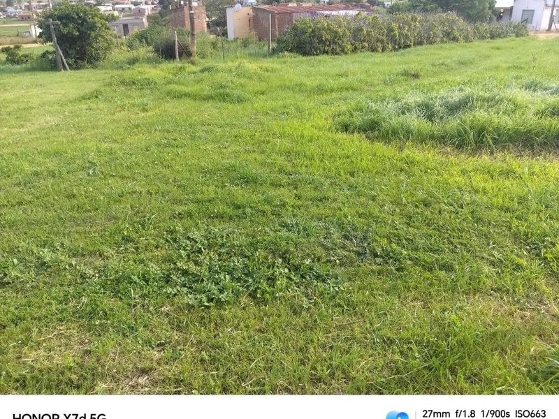 A view of a vacant residential plot featuring a grass lawn and perimeter fencing, situated in a suburban neighborhood setting.