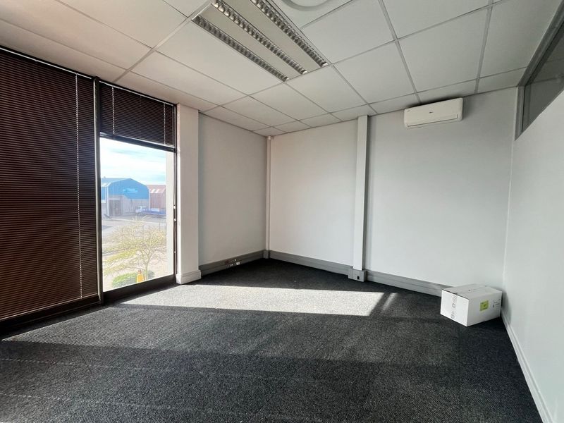 A clean, empty office space featuring a suspended ceiling, recessed lighting, wall-mounted air conditioning, and large windows with venetian blinds.