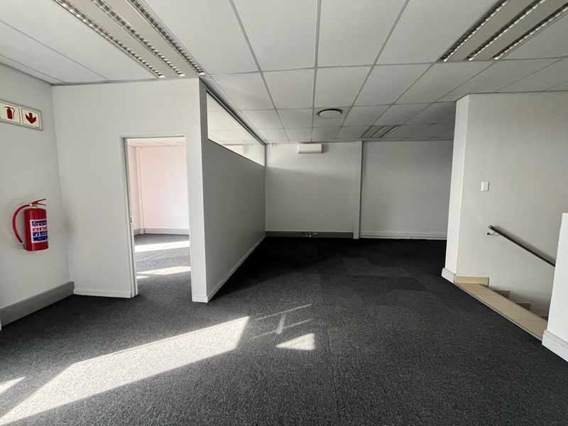 A spacious, empty industrial office area featuring carpeted floors, a suspended ceiling with recessed lighting, and a wall-mounted air conditioning unit.