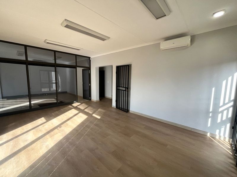 A bright, empty office space featuring laminate flooring, a glass partition wall, security gate, and a wall-mounted air conditioning unit.