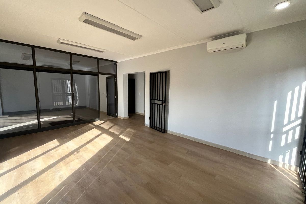 A bright, empty office space featuring laminate flooring, a glass partition wall, security gate, and a wall-mounted air conditioning unit.