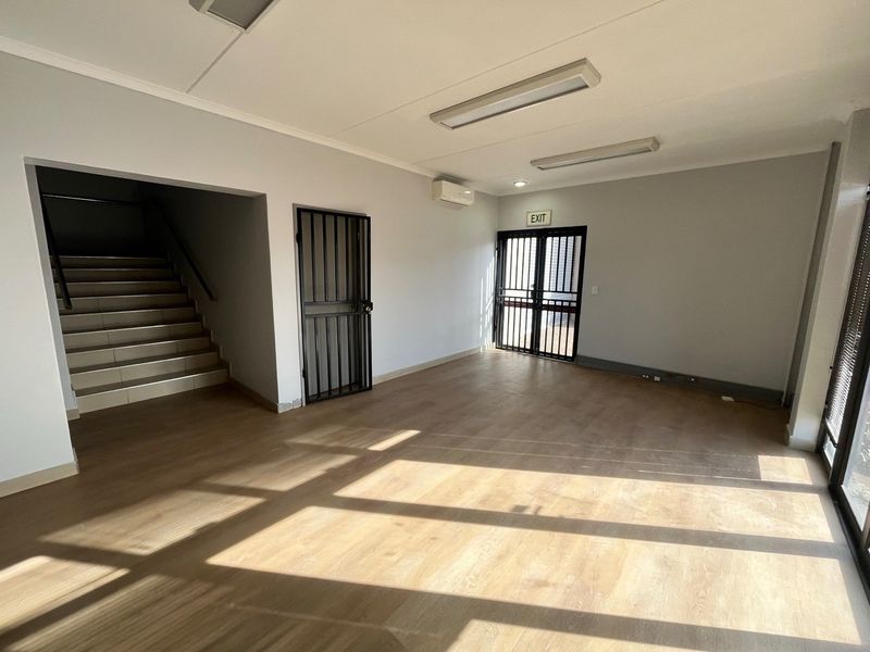 A bright, open-plan office space featuring laminate flooring, a split-system air conditioner, security gates, and direct access to a staircase.