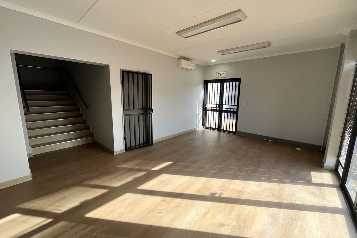 A bright, open-plan office space featuring laminate flooring, a split-system air conditioner, security gates, and direct access to a staircase.