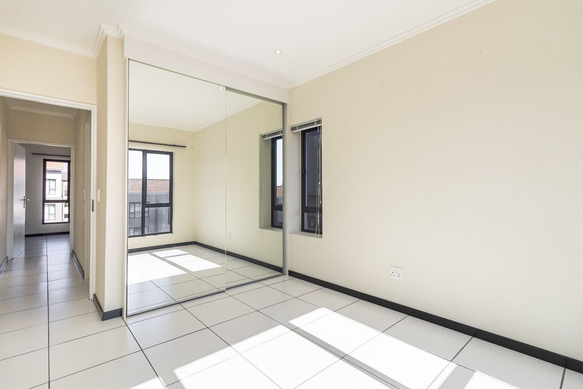 A bright, empty bedroom featuring large built-in mirrored wardrobes, neutral tiled flooring, and modern aluminium window frames.