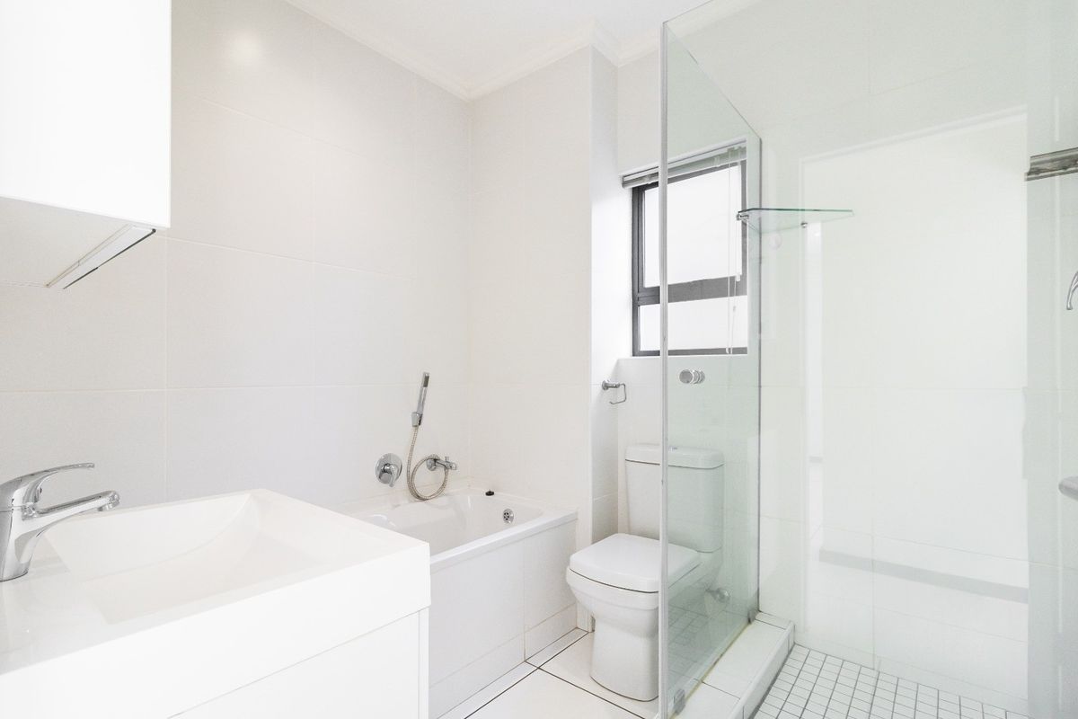 A clean, modern bathroom featuring a built-in vanity, a bathtub, a separate glass-enclosed shower, and a toilet with neutral white tiling throughout.