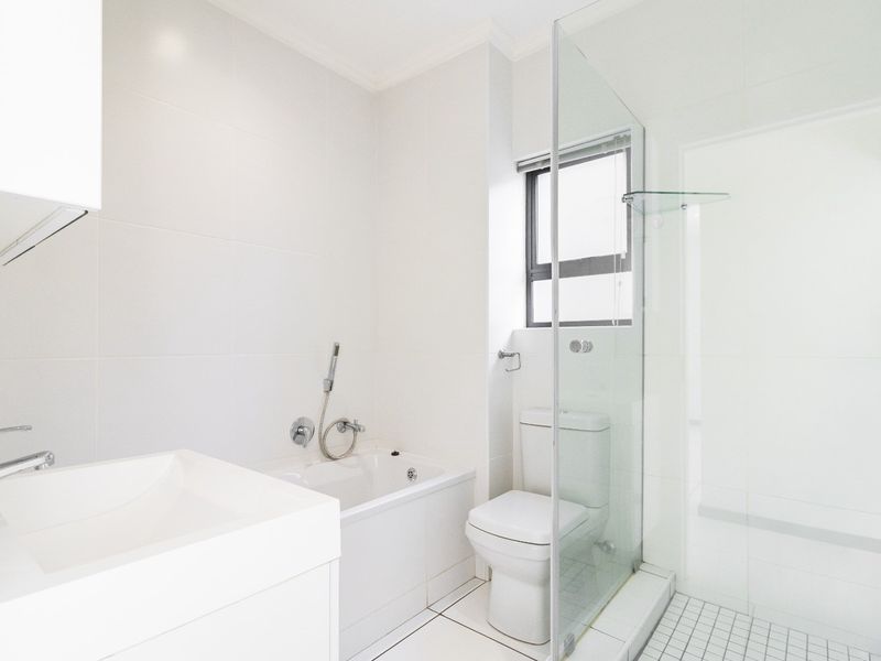 A clean, modern bathroom featuring a built-in vanity, a bathtub, a separate glass-enclosed shower, and a toilet with neutral white tiling throughout.
