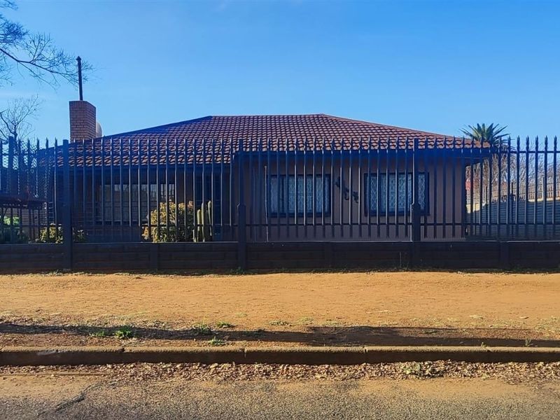 Front elevation of a single-story house featuring a tiled roof, brick chimney, and secure metal perimeter fencing.