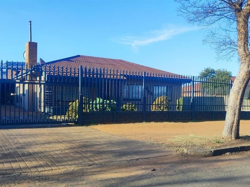 Front exterior view of a single-story house featuring a tiled roof, brick chimney, and secure metal perimeter fencing with a paved driveway.