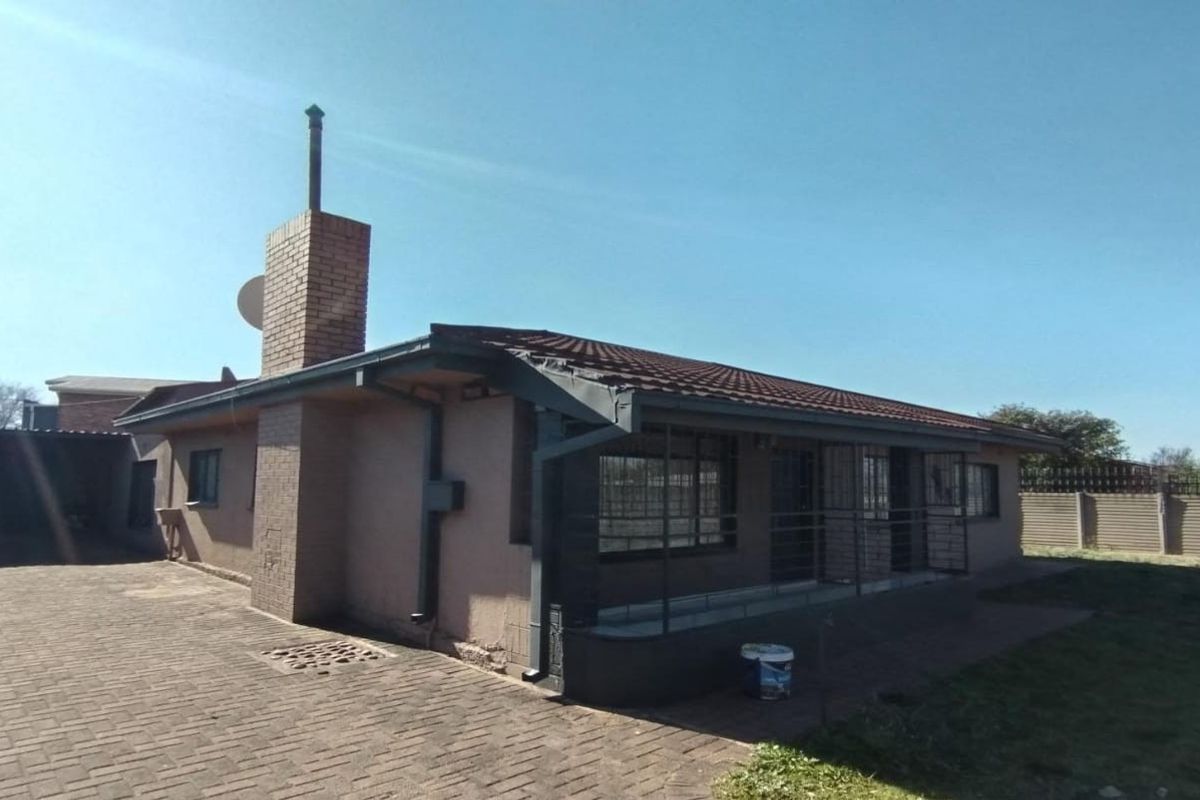 Rear view of a single-story house featuring a brick chimney, tiled roof, paved driveway, and security bars on windows.