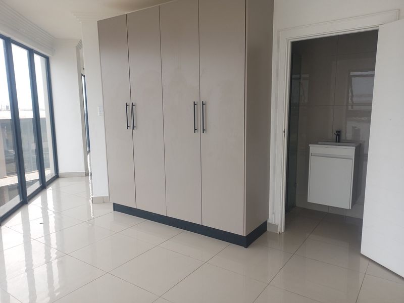A bright, empty bedroom featuring modern built-in cupboards, tiled flooring, and an en-suite bathroom with a vanity unit.