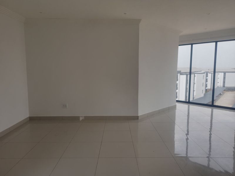 A bright, empty living area featuring polished tiled flooring, recessed lighting, and large floor-to-ceiling windows offering natural light and views of the complex.