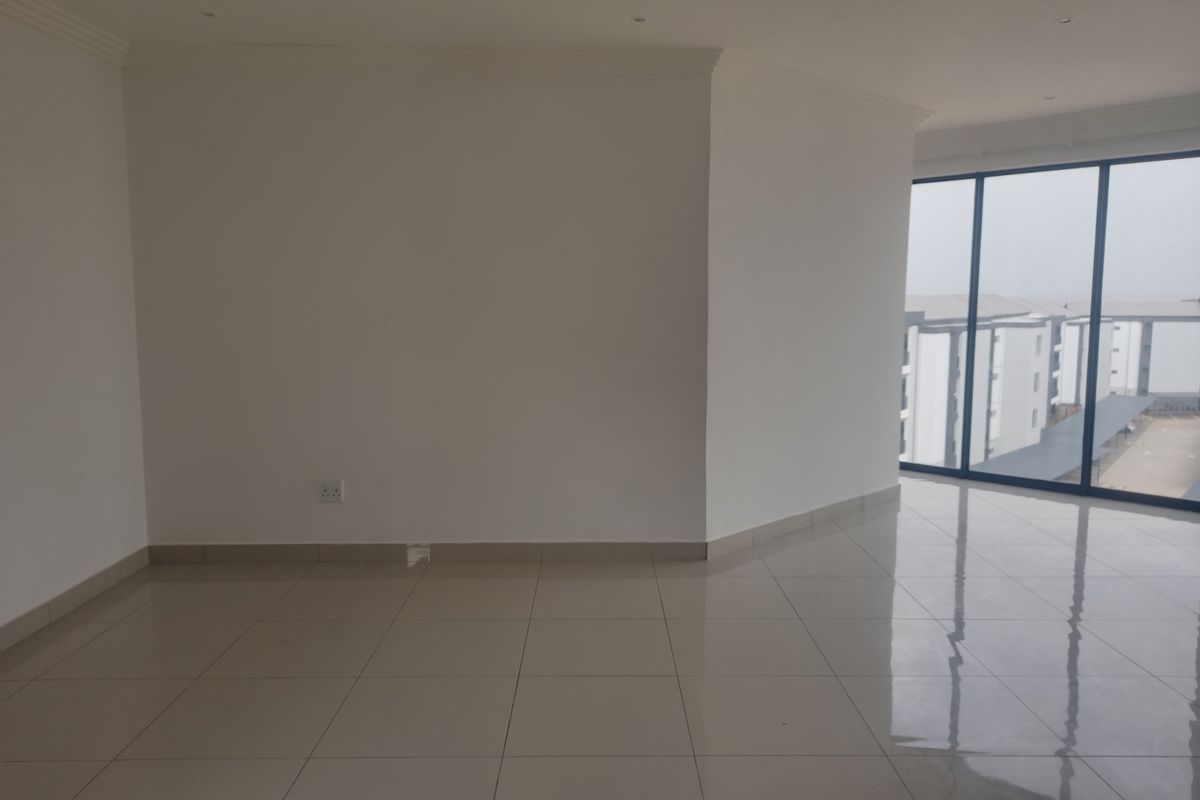 A bright, empty living area featuring polished tiled flooring, recessed lighting, and large floor-to-ceiling windows offering natural light and views of the complex.