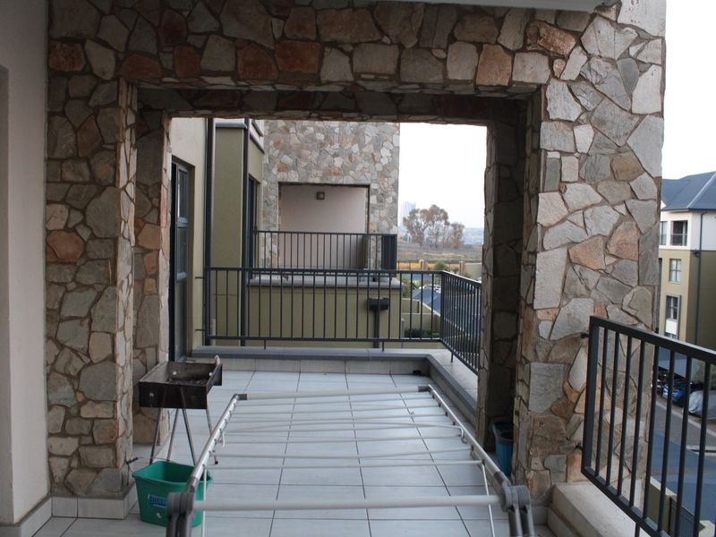 A spacious balcony featuring stone-clad pillars, tiled flooring, and metal balustrades, offering an open-air space with views of the surrounding apartment complex.