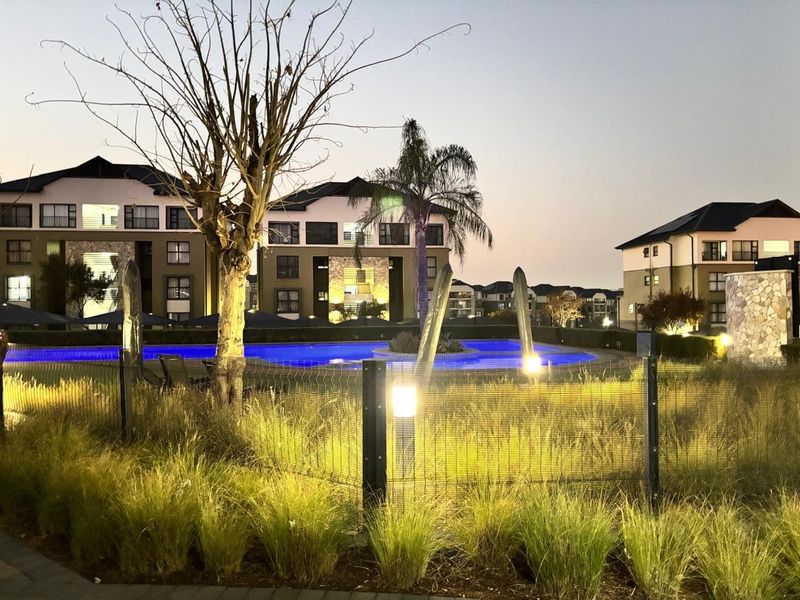 A well-maintained communal swimming pool area within an apartment complex, featuring landscaped gardens, perimeter fencing, and integrated evening lighting.