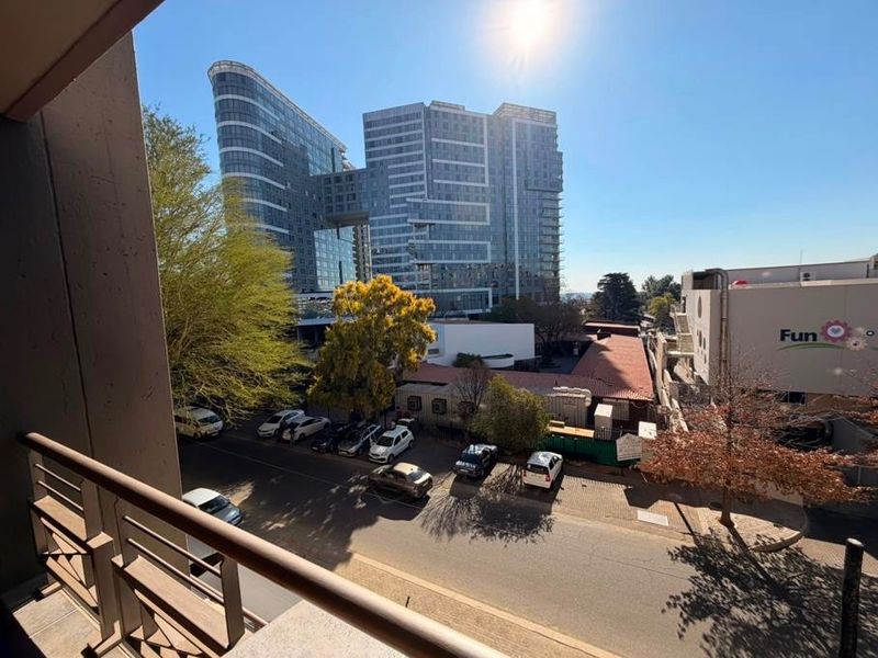 An elevated urban view from a balcony overlooking a street, parking area, and surrounding commercial buildings.