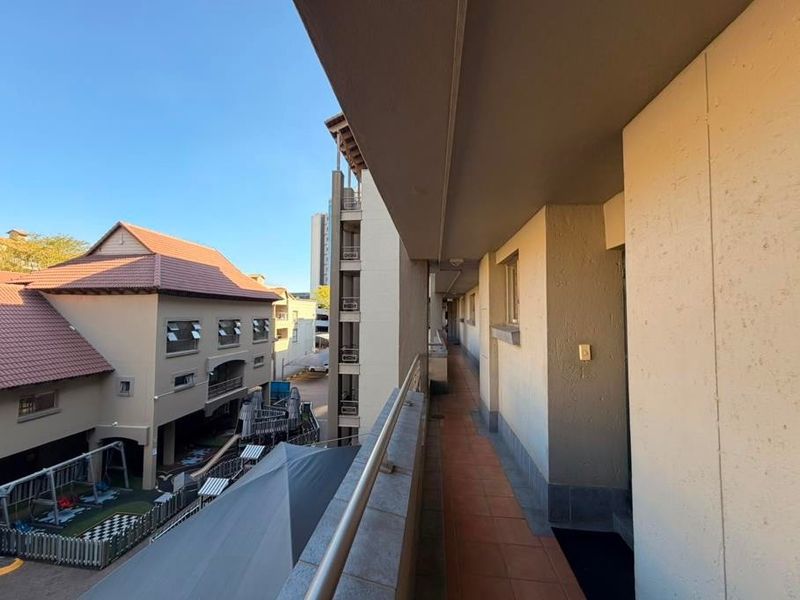 A covered outdoor balcony walkway featuring tiled flooring and a metal balustrade, overlooking the apartment complex grounds.