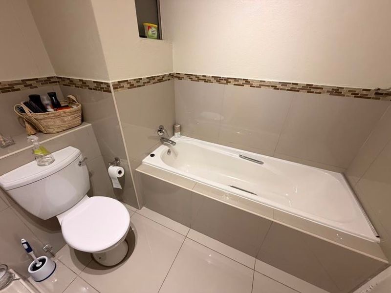 A clean, functional bathroom featuring a built-in bathtub, toilet, and neutral-toned wall and floor tiles with a decorative mosaic border.