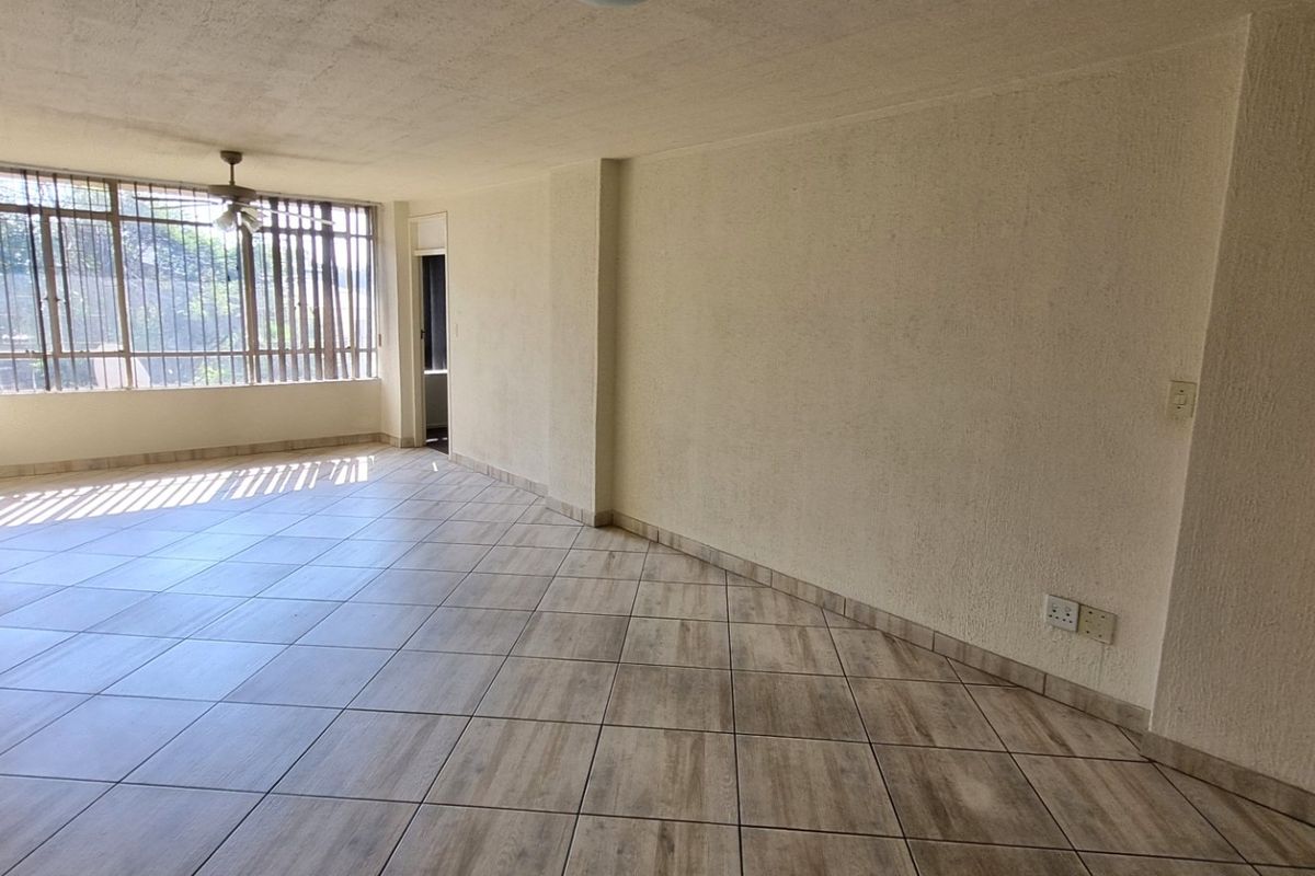 A spacious, empty living area featuring neutral tiled flooring, a ceiling fan, and large windows that provide ample natural light.