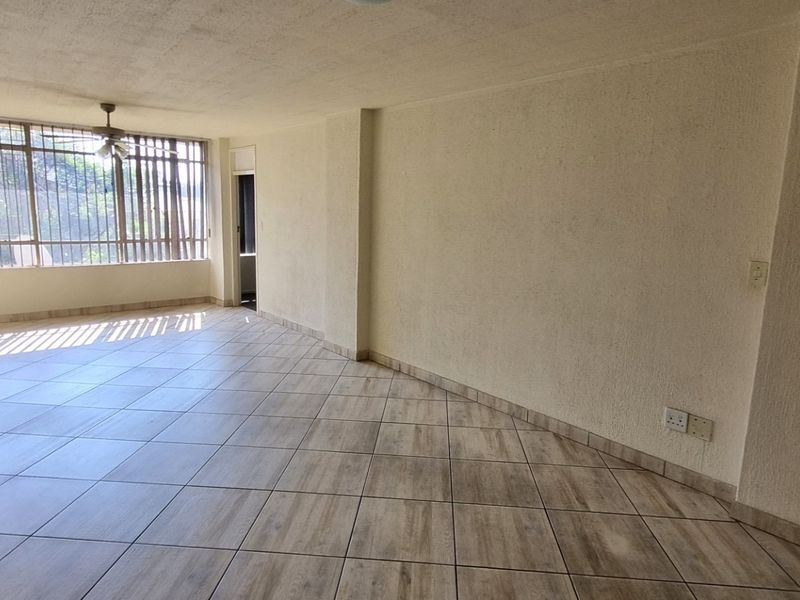A spacious, empty living area featuring neutral tiled flooring, a ceiling fan, and large windows that provide ample natural light.