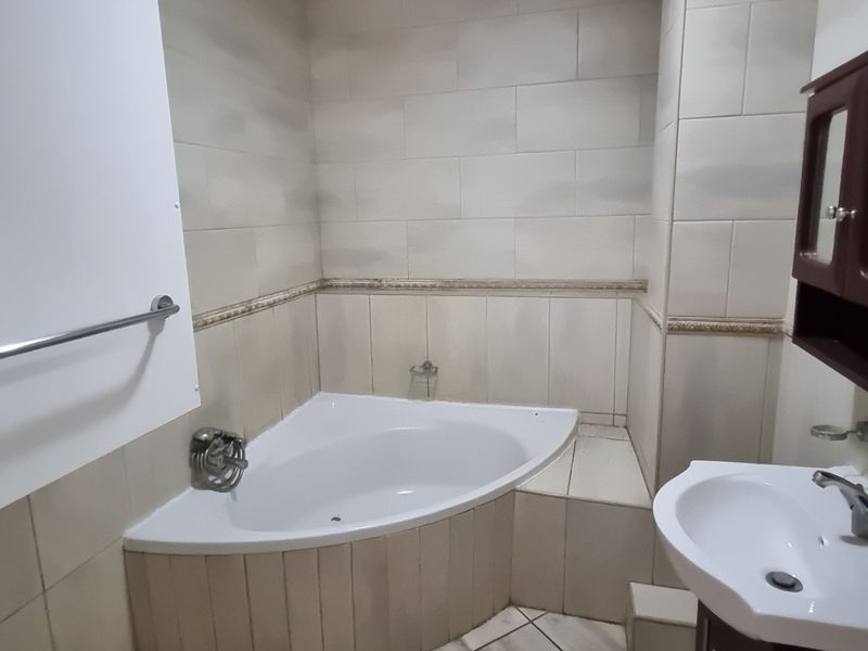 A functional bathroom featuring a corner bathtub, pedestal sink, and wall-mounted storage cabinet with neutral ceramic tiling throughout.