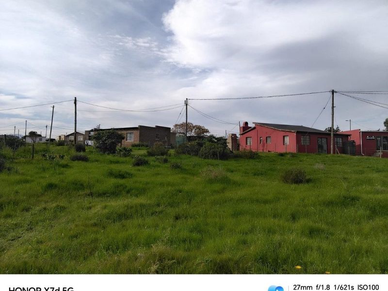 A view of a vacant residential plot featuring grassy terrain with surrounding neighborhood structures and utility infrastructure.