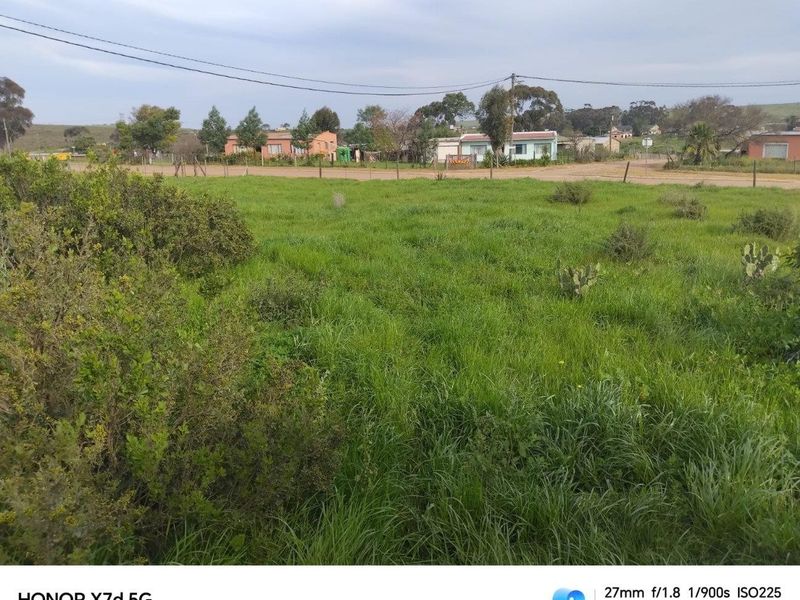 A vacant residential plot featuring natural grass cover, perimeter wire fencing, and overhead power lines, situated in a quiet neighborhood setting.