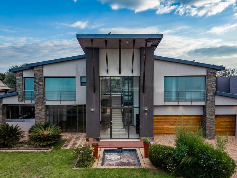 Modern architectural front elevation featuring stone cladding, a prominent glass entrance, double wooden garage doors, and a landscaped front garden with a water feature.
