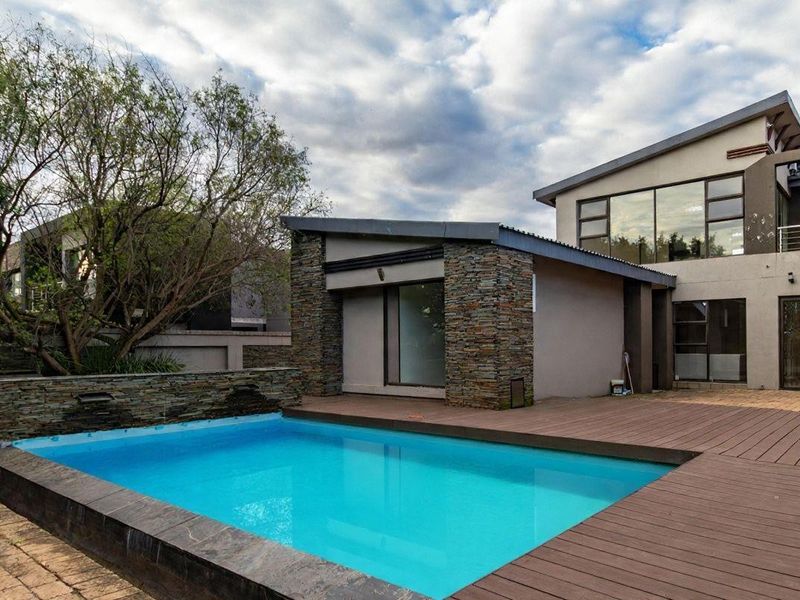 A modern outdoor pool area featuring a swimming pool with a stone-clad water feature, surrounded by timber decking and contemporary architectural building facades.