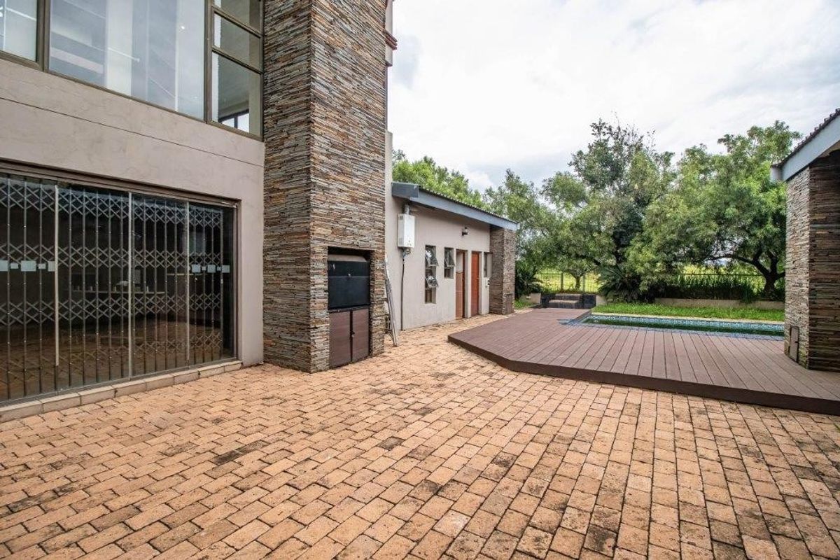 Rear exterior view featuring a paved patio, built-in braai, timber decking surrounding a swimming pool, and stone-clad architectural detailing.
