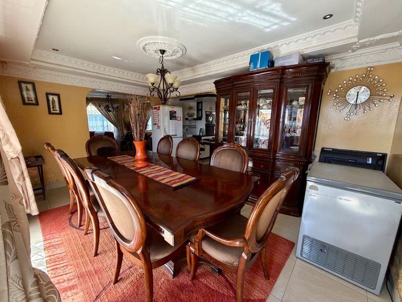A formal dining room featuring ornate ceiling cornicing, a decorative ceiling rose, and tiled flooring, with an open-plan flow into the adjacent kitchen area.