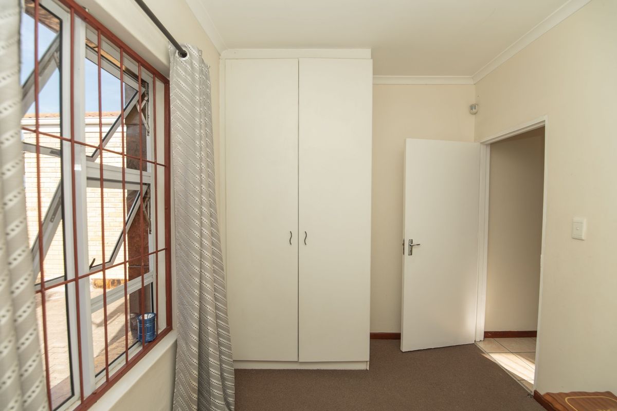 A clean, empty bedroom featuring a built-in wardrobe, carpeted flooring, and a window fitted with security bars.