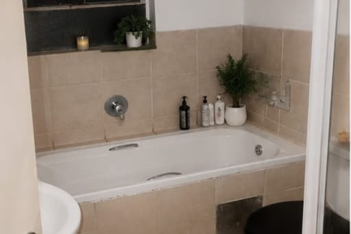 A compact bathroom featuring a built-in bathtub with shower mixer, tiled walls, and a standard toilet, requiring some maintenance and cosmetic updates.