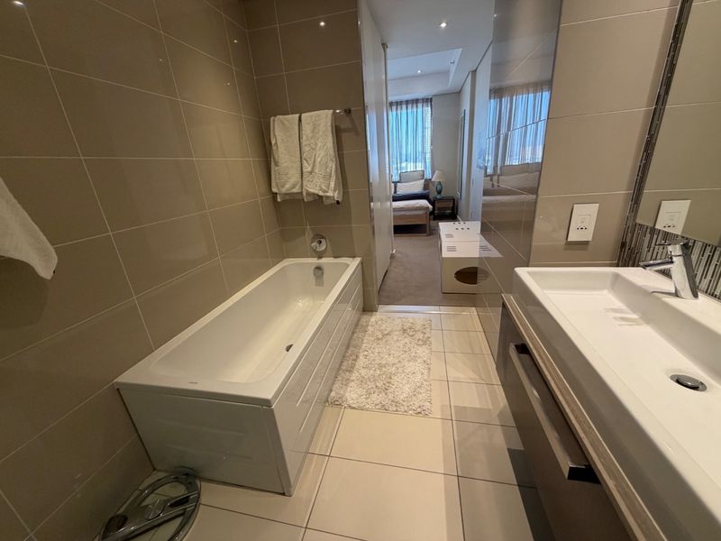 Modern bathroom featuring a built-in bathtub, vanity basin, and neutral floor-to-ceiling tiling, with a view leading into the adjacent bedroom.