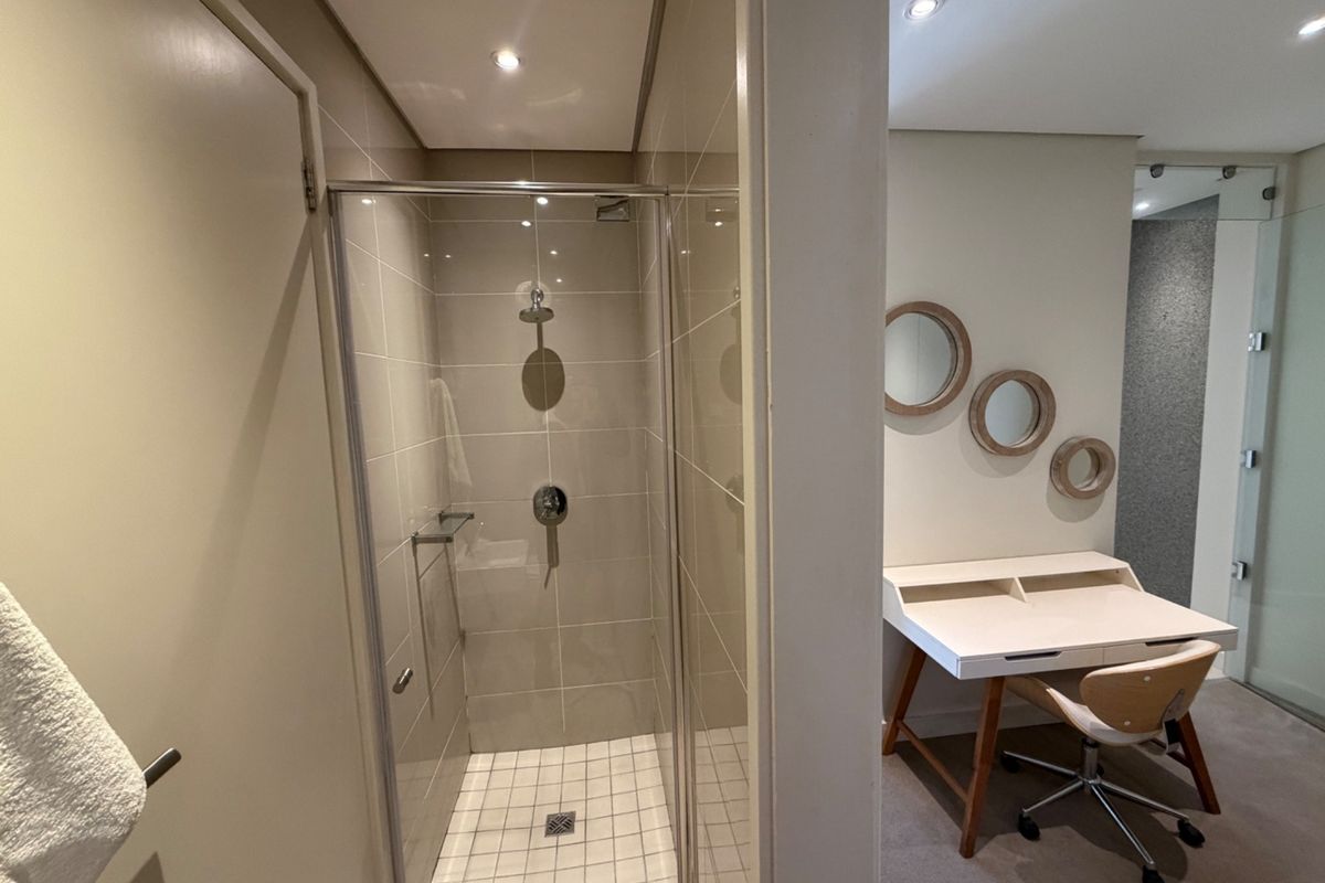 A modern bathroom featuring a glass-enclosed shower with neutral wall tiling and recessed lighting, adjacent to a bedroom workspace area.