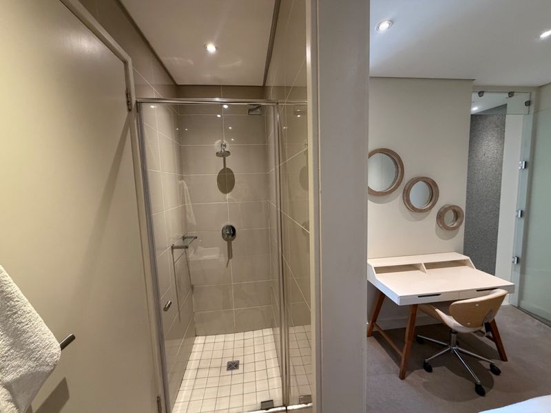 A modern bathroom featuring a glass-enclosed shower with neutral wall tiling and recessed lighting, adjacent to a bedroom workspace area.