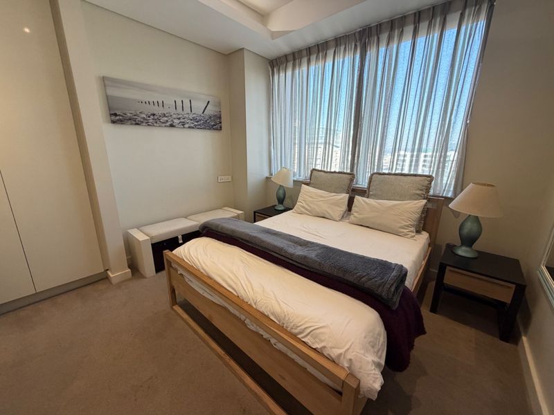 A well-maintained bedroom featuring neutral carpeting, built-in wardrobes, and a large window with sheer curtains allowing for ample natural light.