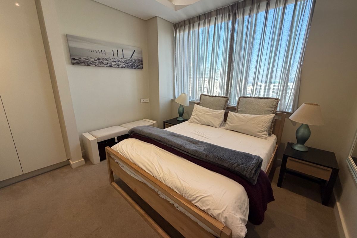 A well-maintained bedroom featuring neutral carpeting, built-in wardrobes, and a large window with sheer curtains allowing for ample natural light.