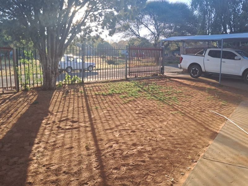 Front yard area featuring a metal security gate, carport structure, and concrete walkway with sparse lawn coverage.