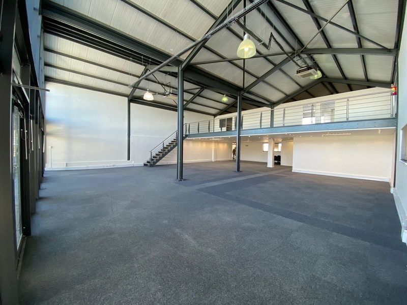 Spacious open-plan office featuring a mezzanine level, industrial-style exposed steel trusses, carpeted flooring, and large glass windows providing ample natural light.