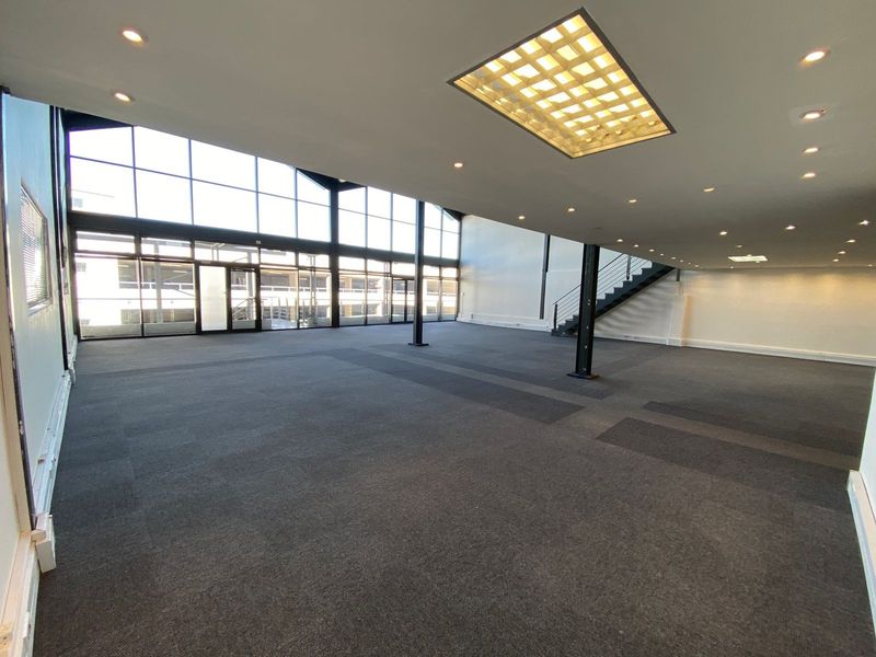 Spacious, open-plan office area featuring high ceilings, large windows for natural light, carpeted flooring, and a modern staircase leading to an upper level.