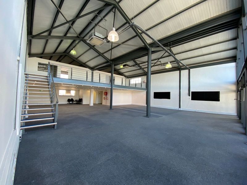 Spacious open-plan office area featuring a mezzanine level, exposed industrial steel trusses, modern pendant lighting, and a metal staircase.
