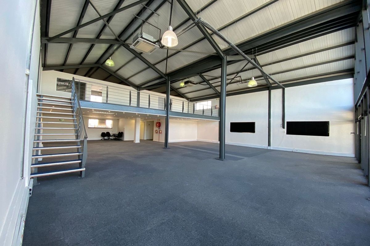 Spacious open-plan office area featuring a mezzanine level, exposed industrial steel trusses, modern pendant lighting, and a metal staircase.